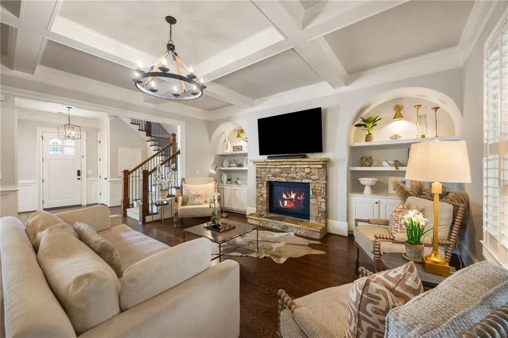 Living room featuring a stone fireplace with hearth, coffered ceiling, hardwood flooring, and built-in arched shelving with cabinetry