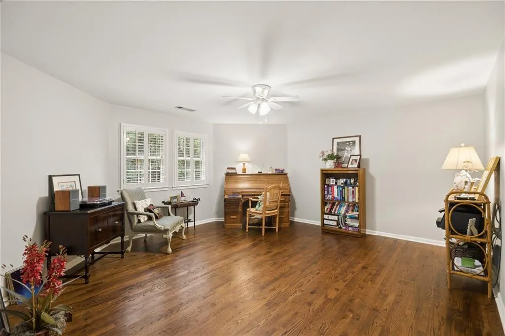 Spacious room featuring hardwood flooring, light gray walls, multiple windows with white shutters, and a ceiling fan