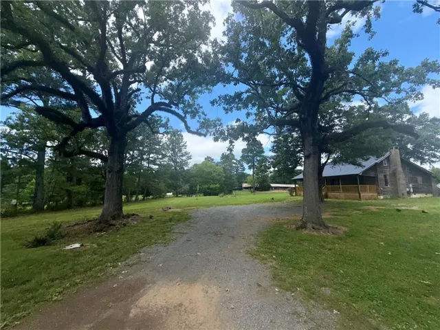 Gravel driveway framed by mature trees, leading to a cabin-style property with a stone chimney and covered porch