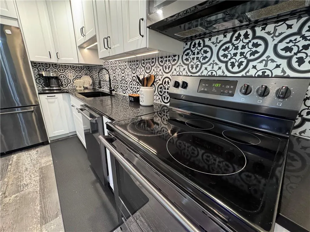 Kitchen with stainless steel appliances, range hood, tasteful backsplash, white cabinetry, and dark stone counters