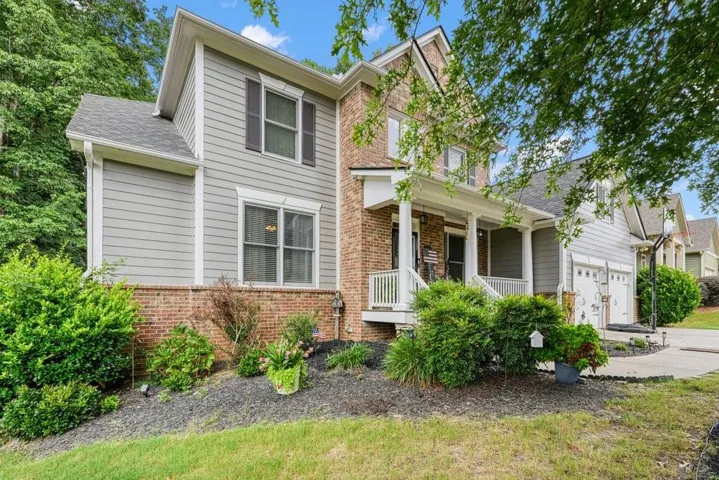Traditional two-story residence featuring a brick facade, siding accents, a covered front porch with white railings, a two-car garage, and mature landscaping
