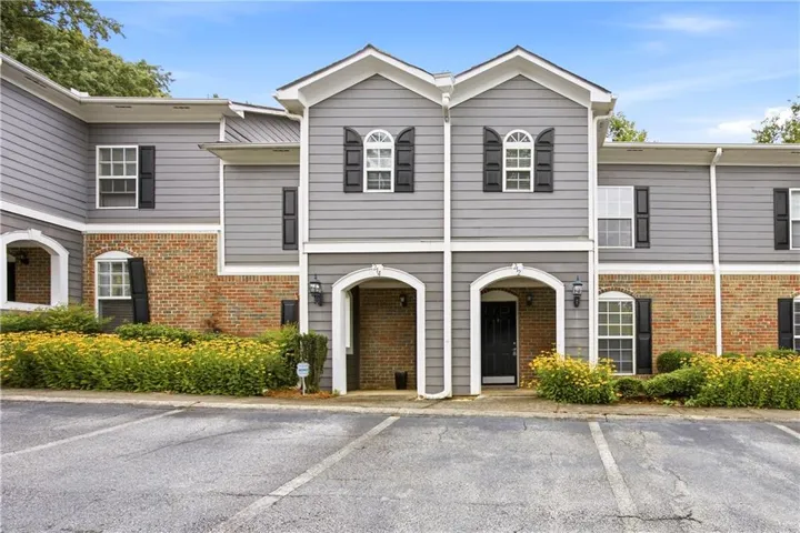 Exterior featuring gray siding, brick accents, arched entryways, black shutters, and mature landscaping with yellow blooms
