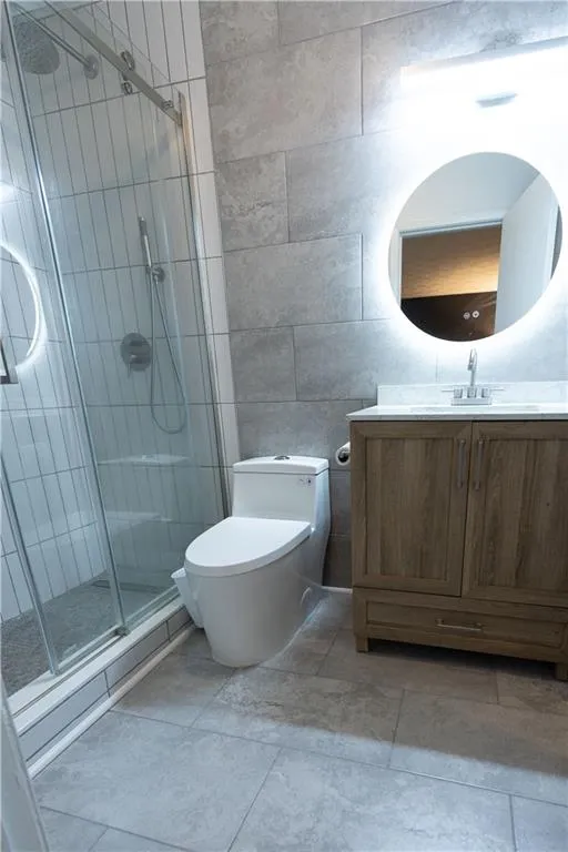Bathroom featuring a glass-enclosed shower with vertical subway tile, a vanity with a light wood-finish cabinet, and a back-lit circular mirror