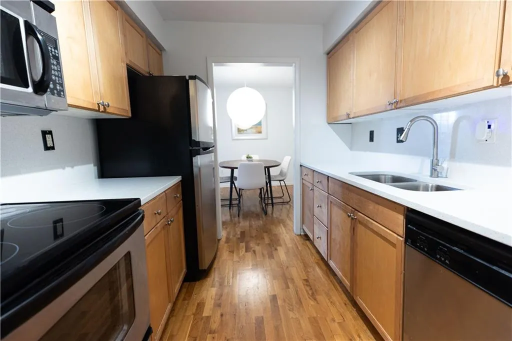 Galley kitchen featuring wood-finish flooring, light wood cabinetry, white countertops, and stainless steel appliances