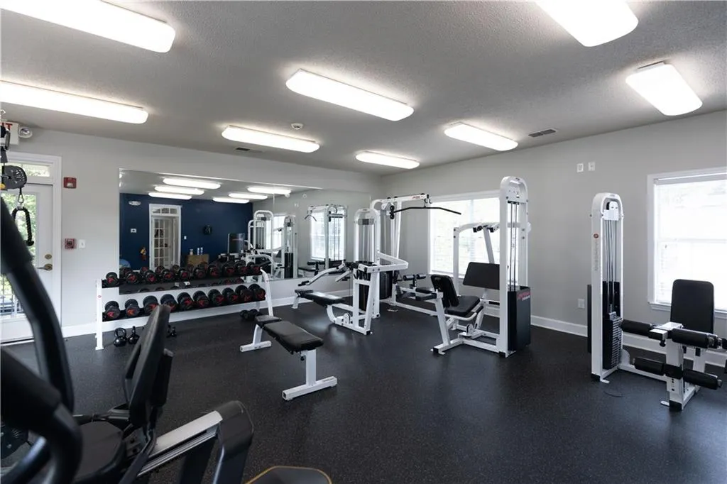 Dedicated fitness space featuring ample natural light from multiple windows, overhead fluorescent lighting, durable black rubber flooring, a large wall-mounted mirror, and a variety of built-in weight machines and free weights