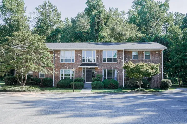 Brick exterior with multiple windows, mature landscaping, and a paved parking area