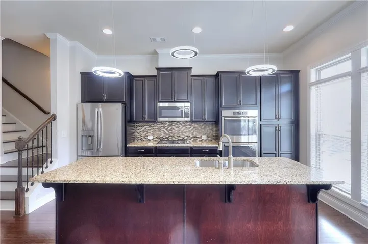 Kitchen featuring an island with a built-in sink, stainless steel appliances, dark cabinetry, and a mosaic tile backsplash