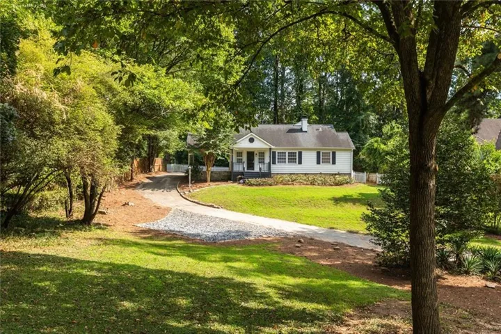 White exterior with dark shutters, covered front porch, gravel driveway, mature trees, and a sloped, manicured lawn