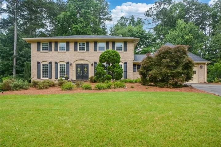 Brick facade residence featuring a two-story design with a double-door entry, black window shutters, an attached two-car garage, and a lush lawn with mature landscaping