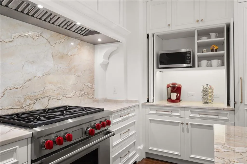 Kitchen featuring custom exhaust hood, white cabinetry, appliances with stainless steel finishes, light stone countertops, and open shelves Kitchen featuring custom exhaust hood, white cabinetry, appliances with stainless steel finishes, light stone countertops, and open shelves