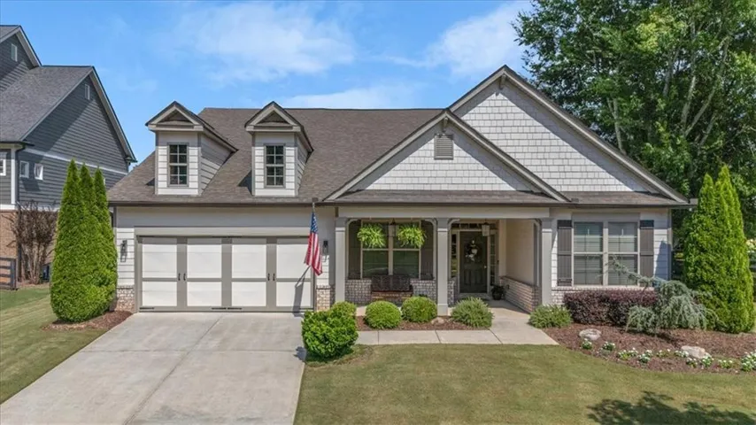 Ranch-style residence featuring a covered front porch, a two-car garage, dormer windows, a shingled roof, and stone-accented siding