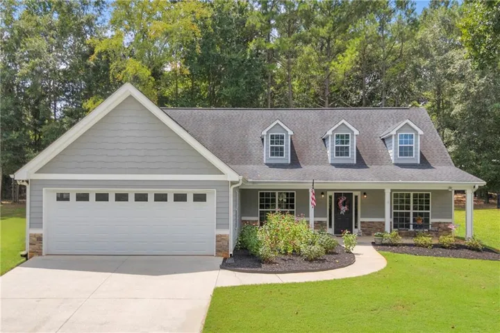Grey siding exterior with stone accents, a covered front porch, a two-car garage with transom windows, a dark shingle roof, and three dormer windows
