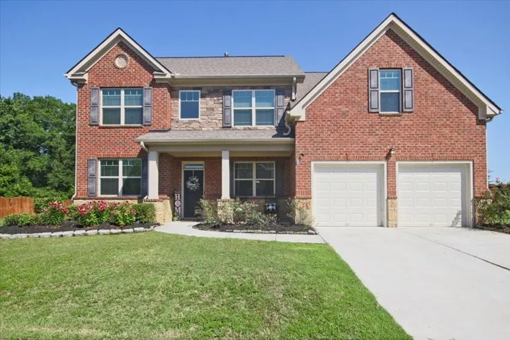 Brick and stone facade featuring a covered front porch with white columns, a two-car garage, manicured lawn, and planted flower beds