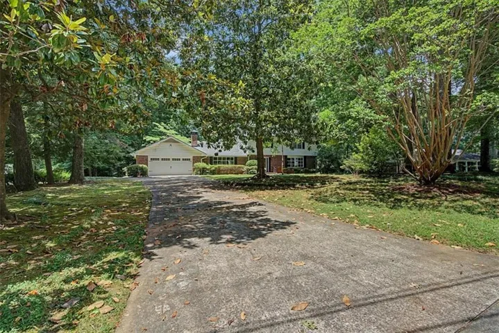 Brick-front residence set back from a concrete driveway, featuring a two-car garage with white paneled doors, expansive front lawn, and mature trees providing abundant shade