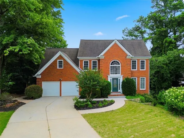 Brick-front residence featuring a two-car garage, arched window, gabled roofline, mature landscaping, and a concrete driveway