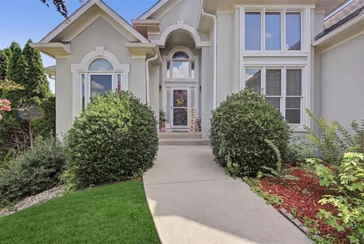 Stucco exterior featuring arched and rectangular windows, a concrete walkway, mature shrubs, and a partial lawn area