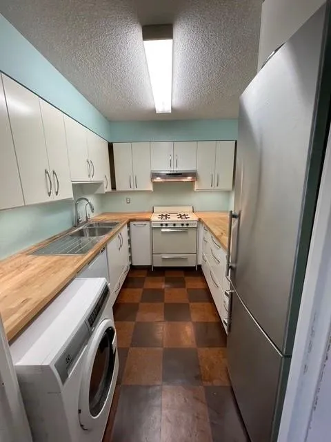 Kitchen with white appliances, a textured ceiling, wooden counters, washer / clothes dryer, and white cabinetry