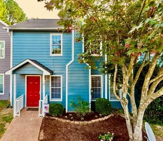 Exterior featuring blue siding, a red front door with a covered entry, a brick paver pathway, and a landscaped garden bed with mature trees
