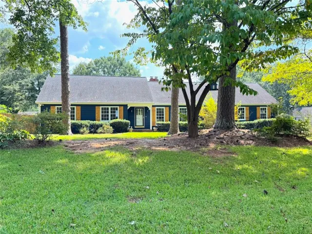 Dark blue siding with light wood-tone shutters, gabled roofline, white trim accents, and mature trees