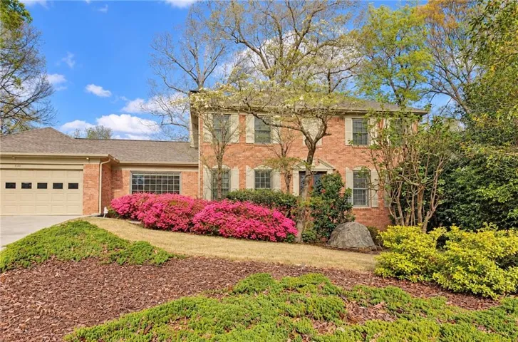 Brick exterior with tan shutters, attached two-car garage, established landscaping, and mature trees