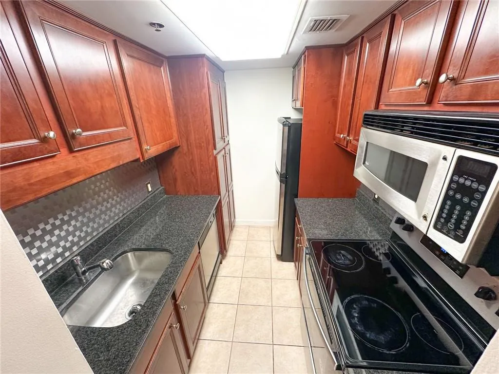 Kitchen with appliances with stainless steel finishes, light tile patterned floors, brown cabinets, dark stone countertops, and decorative backsplash