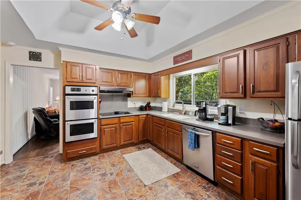 Kitchen with appliances with stainless steel finishes, brown cabinetry, a ceiling fan, dark countertops, and a raised ceiling
