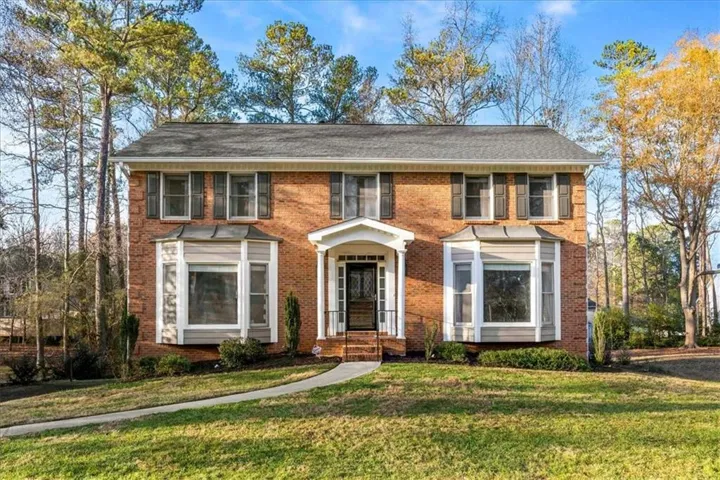 Brick facade residence featuring a covered entry, two bay windows, exterior shutters, and a gray shingle roof