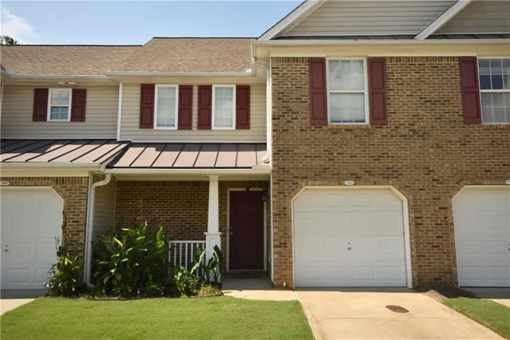 Brick townhome featuring a two-car garage, covered front entry, shuttered windows with white trim, and manicured lawn with mature landscaping