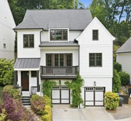 Modern farmhouse residence featuring clean white vertical siding, a multi-level design with a prominent front-facing deck, dual two-car garage bays with paneled doors, metal roof accents, and mature landscaping with established shrubs and trees framing the property