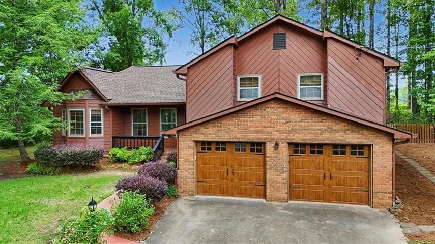 Traditional style residence featuring a reddish-brown exterior with brick accents, a two-car garage with wood-finish doors, a shingle roof, and mature landscaping