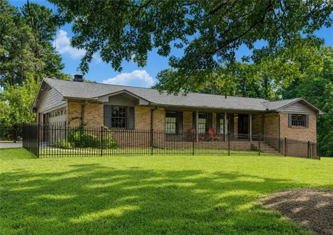 Brick exterior ranch-style home featuring a covered front porch, an attached garage, a black metal fence, and mature trees surrounding the property