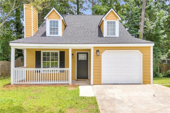 Gable roofline with dormer windows, covered front porch with white railing, yellow siding exterior, attached garage, and a concrete driveway