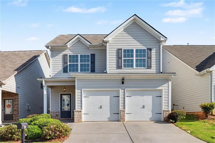 Light gray siding exterior with dark shutters, covered front porch, two-car garage, and established landscaping