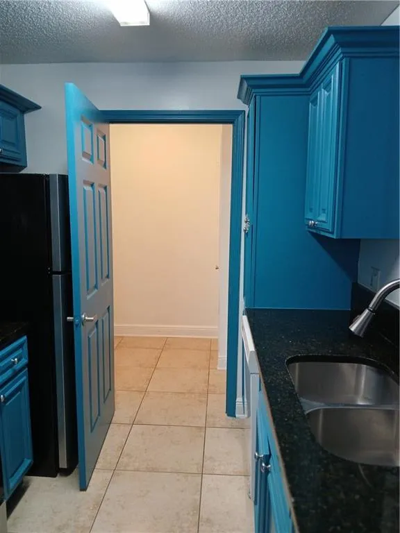 Kitchen featuring blue cabinets, refrigerator, a textured ceiling, light tile patterned floors, and dark stone counters