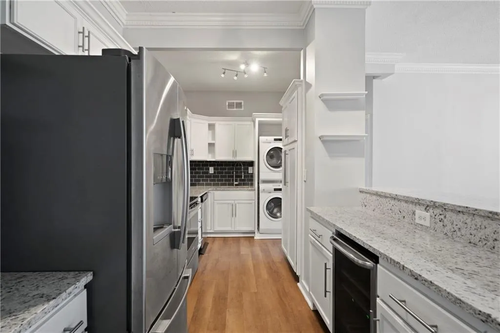 Kitchen with hardwood / wood-style flooring, stainless steel fridge with ice dispenser, white cabinets, stacked washing maching and dryer, and light stone countertops