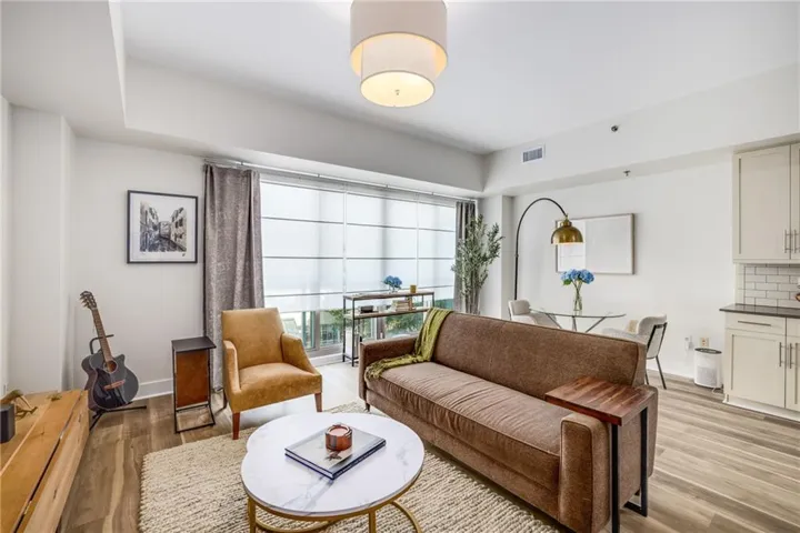 Bright open-concept living area with wood-finish flooring, a contemporary pendant light fixture, and floor-to-ceiling windows with roller shades