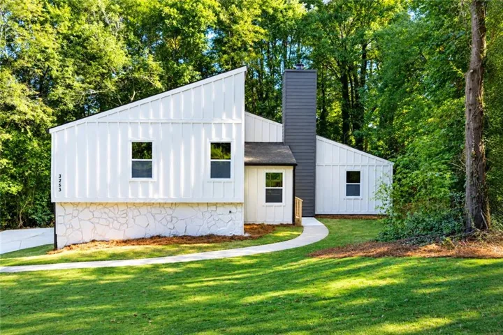 Modern exterior with white vertical siding, a dark gray chimney, multiple windows, a curved walkway, and a natural treeline backdrop