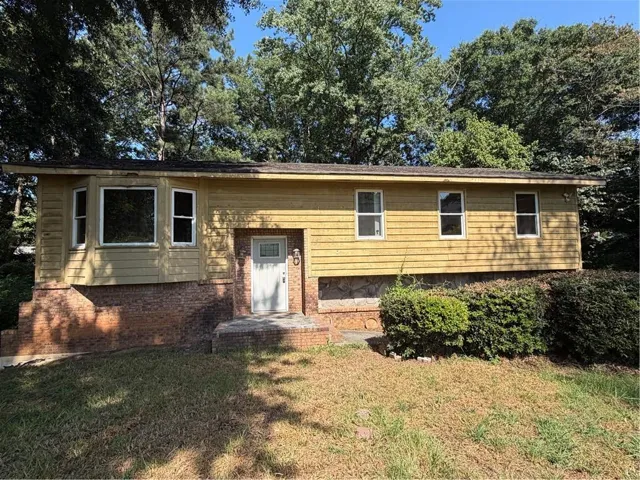 Yellow siding exterior with a white front door, brick foundation, and established perimeter shrubs