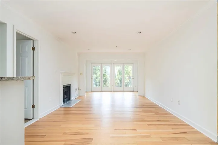 Spacious room featuring light wood-finish flooring, a white mantel fireplace with a black firebox, and French doors with transoms leading to a balcony