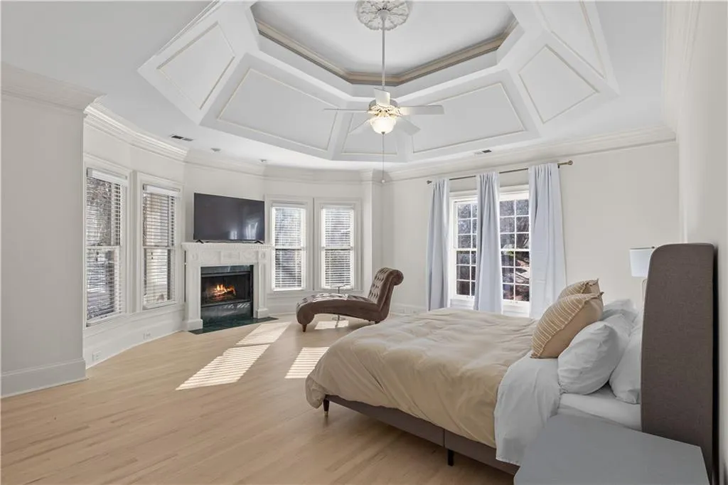 Bedroom featuring light wood-type flooring, a high end fireplace, ornamental molding, a ceiling fan, and a raised ceiling