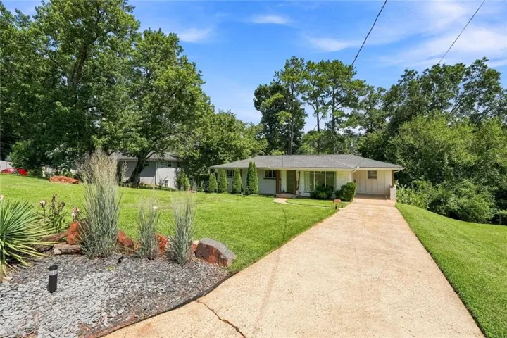 Ranch-style exterior with a concrete driveway, mature trees, and landscaping featuring ornamental grasses and decorative rocks