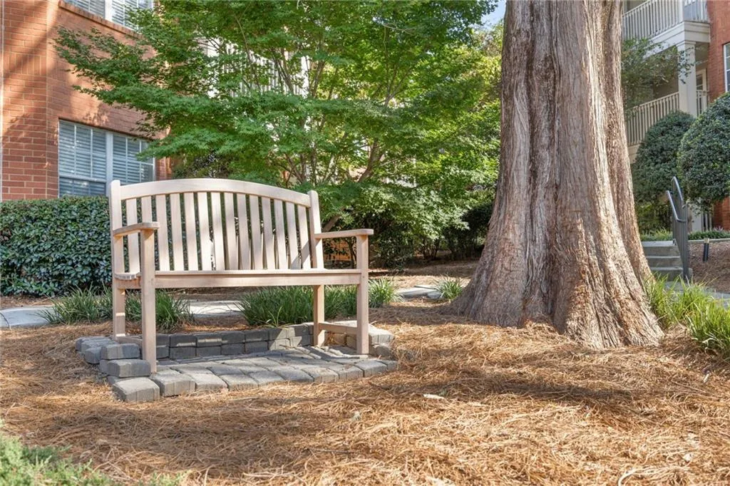 Serene backyard retreat featuring a curved wooden bench nestled beneath a mature shade tree, mulched ground cover, and brick residential architecture visible in the background