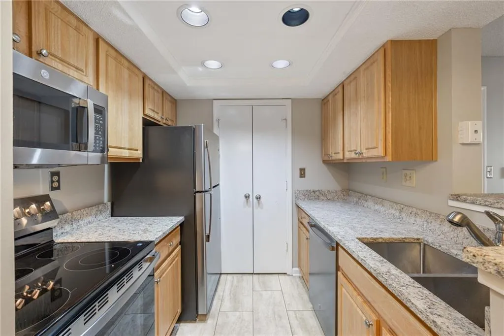 Galley kitchen featuring wood-finish cabinetry, granite countertops, stainless steel appliances including a built-in microwave and electric range, undermount sink, recessed lighting, and tile flooring throughout