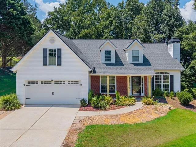 Traditional-style exterior with white siding and brick accents, featuring a two-car garage, multiple dormer windows, and a paved driveway