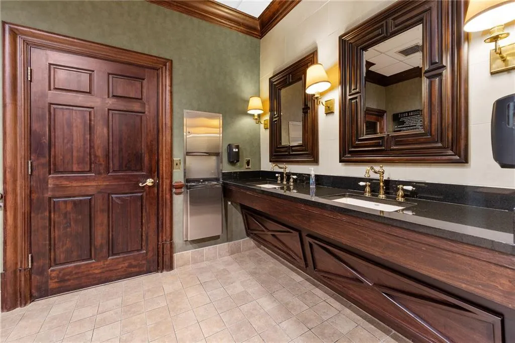 Hardwood door with panel detailing, dual vanity with dark countertop, integrated sinks with gold-finish faucets, decorative wood-framed mirrors, and wall-mounted light fixtures