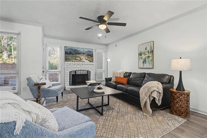 Wood-finish flooring, white brick fireplace, multiple windows with blinds, crown molding, and a ceiling fan with integrated lighting