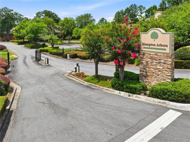 Community entrance featuring a stacked stone monument sign, lush green landscaping, flowering crepe myrtle trees, and an asphalt driveway with entry gates