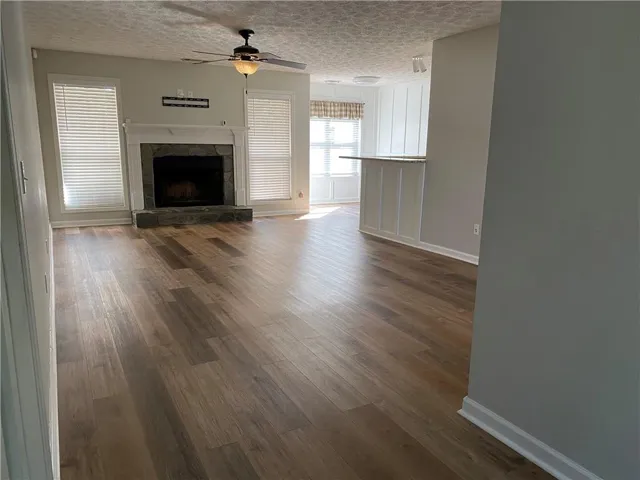 Spacious room with wood-finish flooring, a stone-faced fireplace with a white mantel, and a ceiling fan with integrated lighting