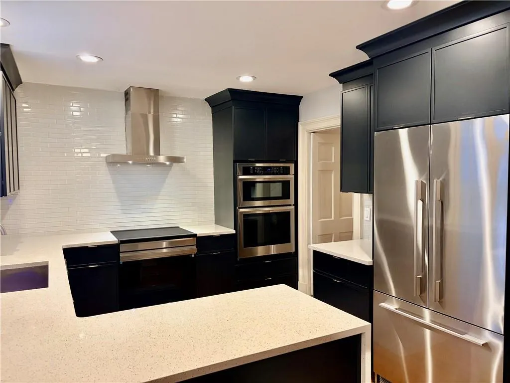 Kitchen featuring dark cabinetry, light-toned countertops, a stainless steel refrigerator, a built-in oven, and a cooktop with a range hood