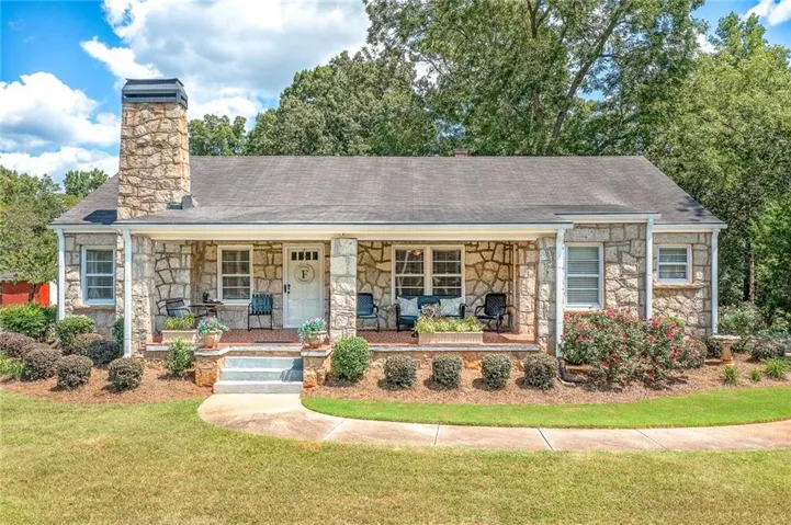 Stone exterior with a full-width covered porch, stone chimney, and landscaping with mature plantings